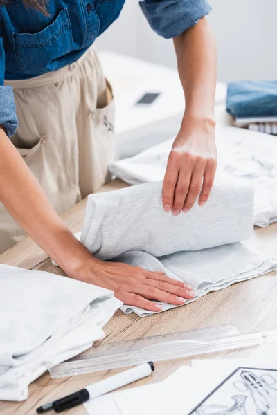 Imagen recortada del diseñador de moda femenino plegable camiseta en la mesa en el estudio de diseño de ropa - foto de stock