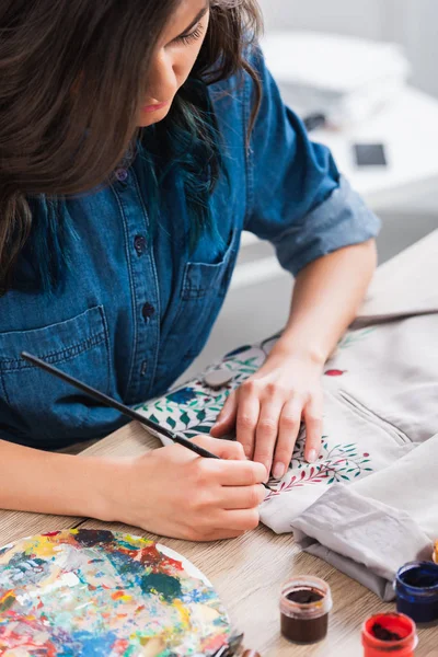 Focused female fashion designer painting on jacket at working table in clothing design studio — Stock Photo
