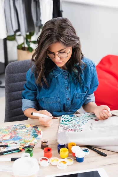Créatrice de mode féminine dans la peinture de lunettes sur veste à la table de travail dans le studio de conception de vêtements — Photo de stock