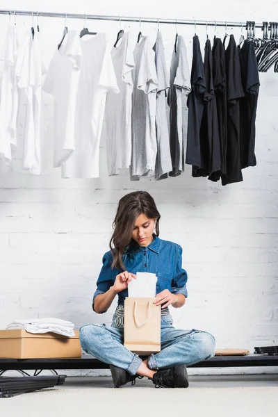 Mujer diseñadora poniendo camiseta blanca en bolsa de papel en estudio de diseño de moda - foto de stock