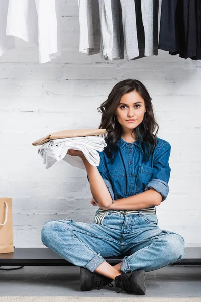 Designer feminino sentado com pilha de t-shirts brancas e pacote de papel em estúdio de design de roupas — Fotografia de Stock