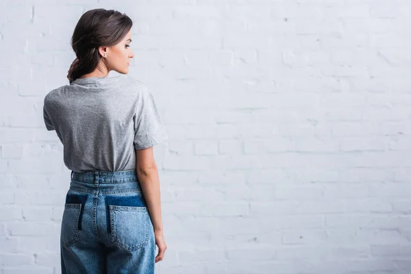 Vista lateral de la mujer joven mostrando la parte posterior vacía de la camiseta gris delante de la pared de ladrillo - foto de stock