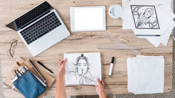 Vista aérea del diseñador femenino que pone la camiseta blanca con la impresión en la mesa con los dispositivos digitales y pinturas - foto de stock