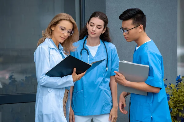 Teacher looking at documents and standing with multicultural students at medical university — Stock Photo