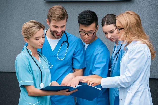 Teacher and multicultural students looking at folder on street near medical university — Stock Photo