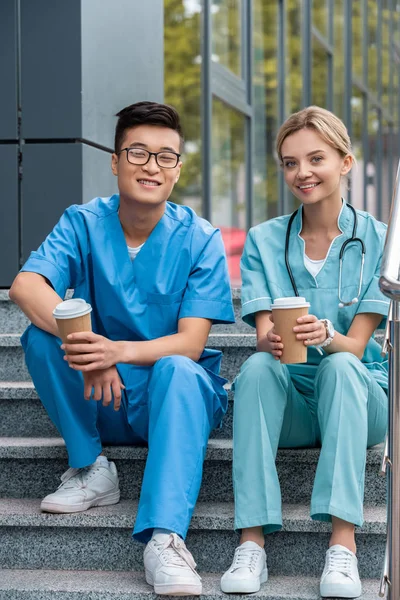 Estudiantes de medicina multicultural sentados en escaleras con tazas de café desechables - foto de stock