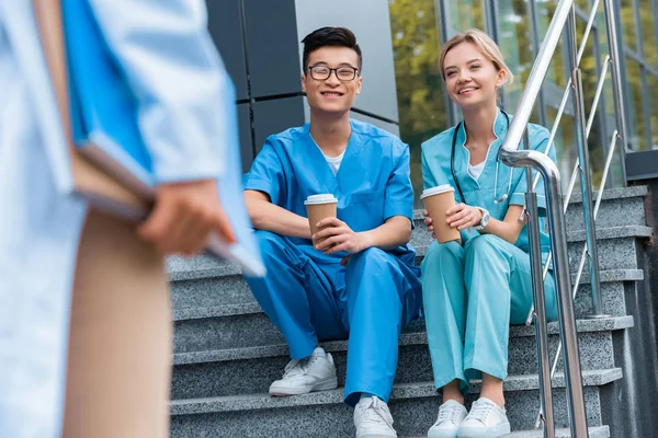 Imagen recortada del profesor mirando a los estudiantes de medicina sonrientes sentados en las escaleras con café para ir - foto de stock