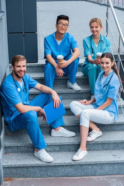Улыбающиеся мультикультурные студенты-медики сидят на лестнице и смотрят в камеру — стоковое фото