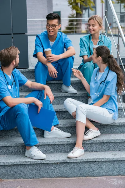 Улыбающиеся мультикультурные студенты-медики разговаривают, сидя на лестнице — стоковое фото
