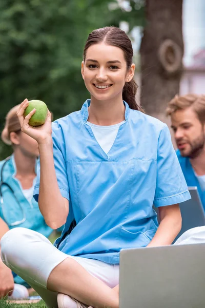 Smiling medical student holding ripe green apple and sitting with laptop — Stock Photo
