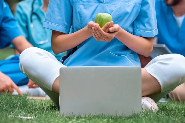 Immagine ritagliata di studente di medicina in possesso di mela verde matura in mano e seduto vicino al computer portatile — Foto stock