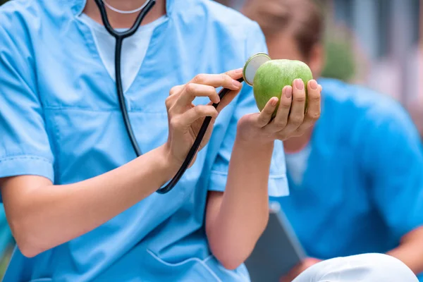 Imagen recortada de estudiante de medicina examinando manzana con estetoscopio - foto de stock