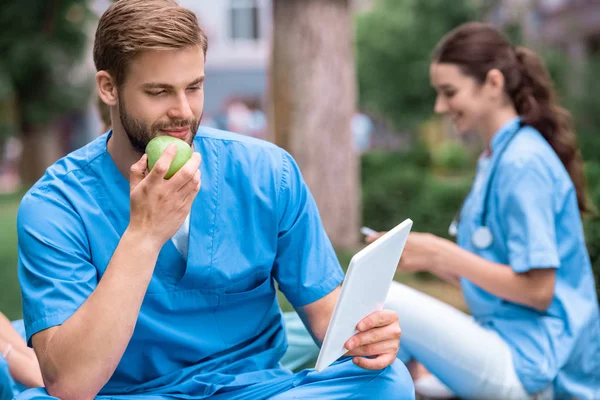 Schöner kaukasischer Medizinstudent isst Apfel und schaut auf Tablet — Stockfoto