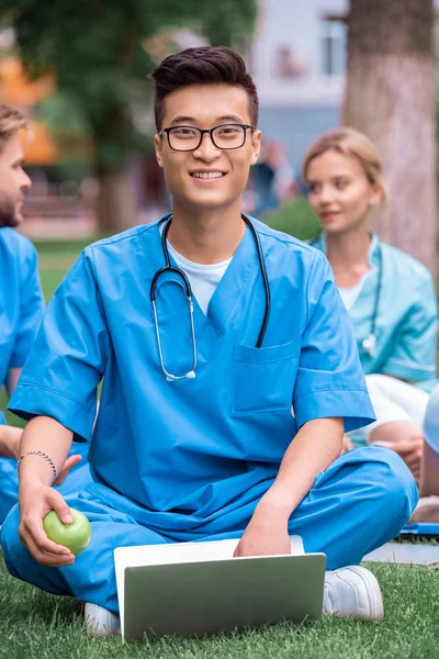 Guapo asiático médico estudiante celebración de manzana con portátil y mirando a cámara - foto de stock