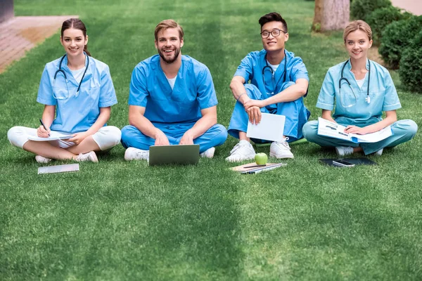 Lächelnde multikulturelle Medizinstudenten, die auf Gras sitzen und in die Kamera schauen — Stockfoto