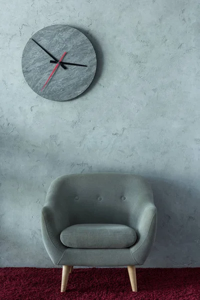 Серый кресло на бордовом ковре возле серой стены в офисе, часы на стене — стоковое фото