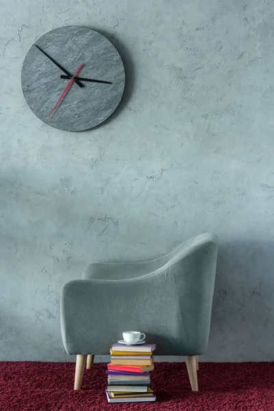 Стопка книг и чашка кофе возле серого кресла в офисе, часы на стене — стоковое фото