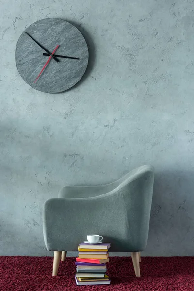 Stack of books and cup of coffee near grey armchair in office, clock on wall — Stock Photo