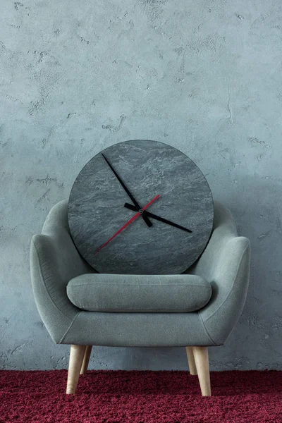 Fauteuil gris avec horloge sur tapis bordeaux près du mur gris dans le bureau — Photo de stock