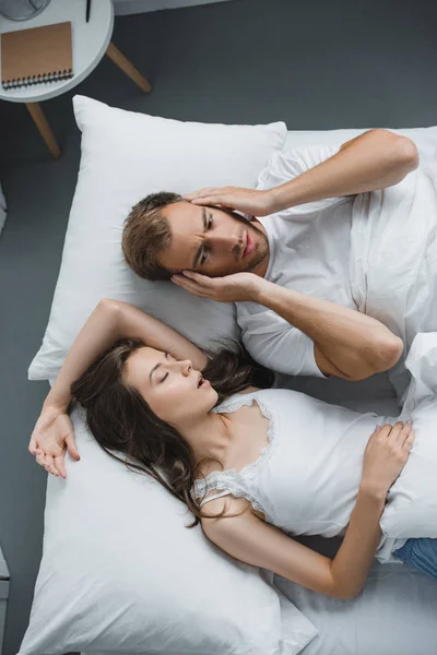 Top view of man closing ears and looking at snoring woman in bed — стоковое фото