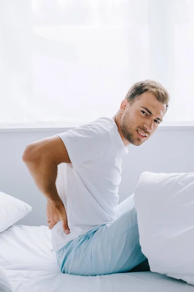 Seitenansicht eines an Rückenschmerzen leidenden Mannes, der im Schlafzimmer in die Kamera schaut — Stockfoto