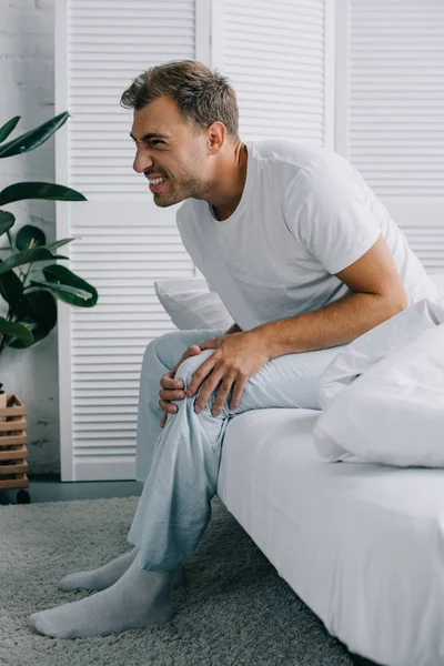 Young man with pain in knee sitting on bed and looking away at home — Stock Photo