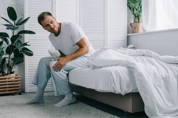 Молодой человек в пижаме сидит на кровати и смотрит в сторону, страдая от боли в колене дома — стоковое фото