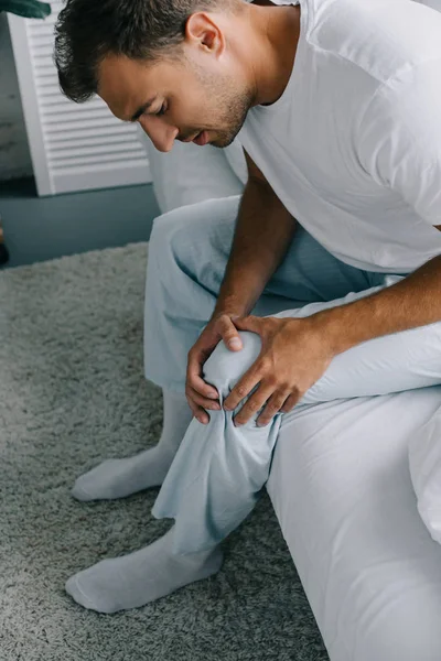 Высокий угол обзора человека в пижаме, страдающего от боли в колене, сидя на кровати — стоковое фото