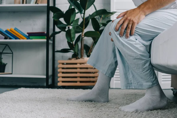 Cropped shot of young man in pajamas suffering from knee pain while sitting on bed — Stock Photo