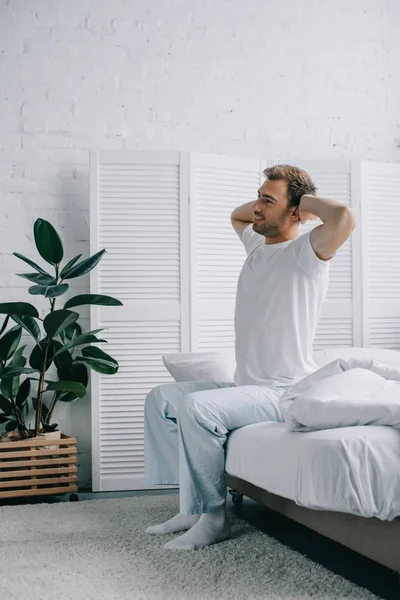 Вид збоку щасливого молодого чоловіка в піжамі розтягується, сидячи на ліжку — стокове фото