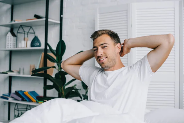 Schöner junger Mann streckt die Hände aus und lächelt in die Kamera, während er morgens auf dem Bett sitzt — Stockfoto