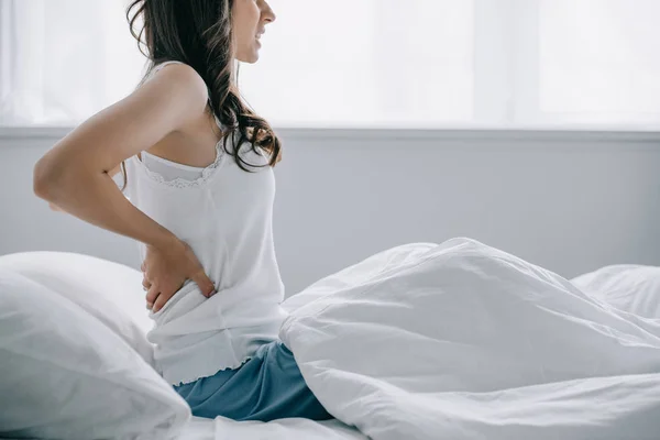 Cropped shot of young woman in pajamas suffering from back pain while sitting on bed — Stock Photo