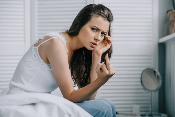 Junge Frau mit Kopfschmerzen gibt den Finger und blickt im Schlafzimmer in die Kamera — Stockfoto