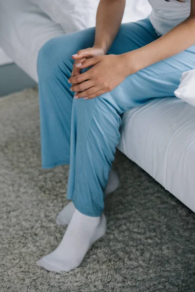 Cropped shot of woman in pajamas sitting on bed and suffering from pain in knee — Stock Photo