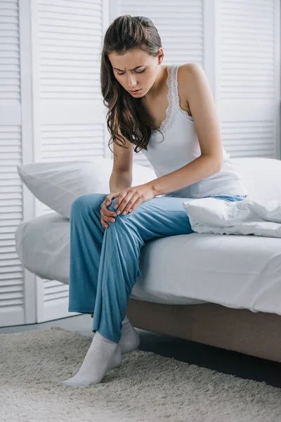 Attractive young woman in pajamas suffering from pain in knee while sitting on bed at home — Stock Photo