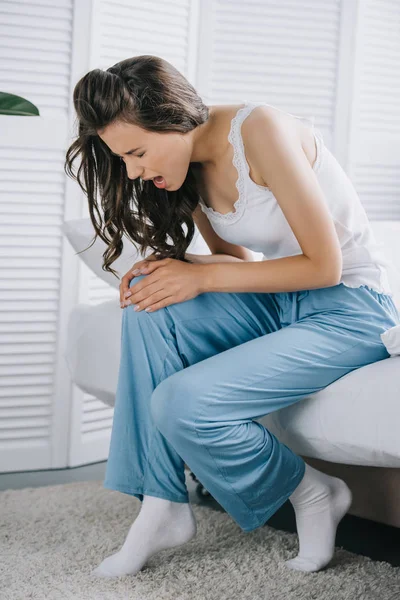 Young woman in pajamas suffering from knee pain while sitting on bed at home — Stock Photo