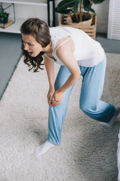 Vue grand angle de la jeune femme en pyjama souffrant de douleur au genou à la maison — Photo de stock