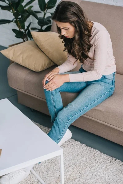 Hochwinkelaufnahme einer Frau mit Schmerzen im Knie, die zu Hause auf dem Sofa sitzt — Stockfoto