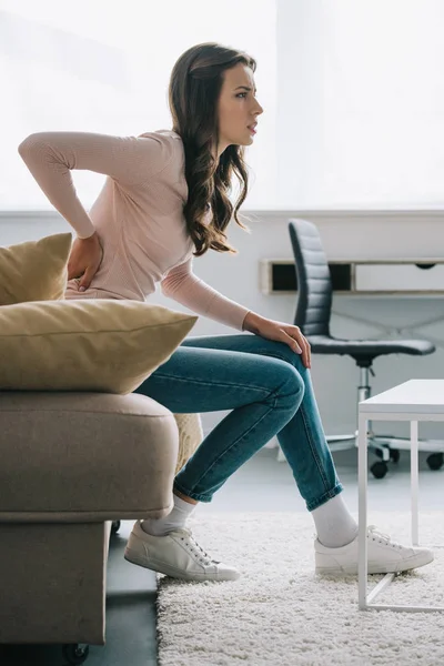 Side view of young woman sitting on sofa and suffering from back pain — Stock Photo