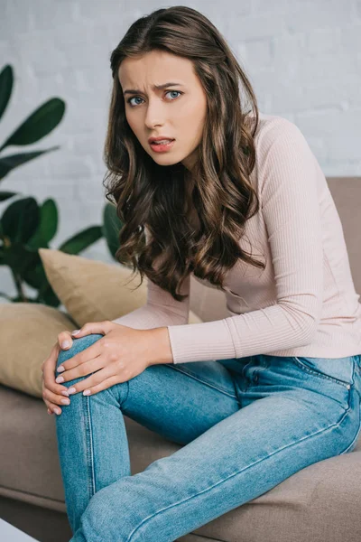 Young woman with pain in knee sitting on sofa and looking at camera — Stock Photo