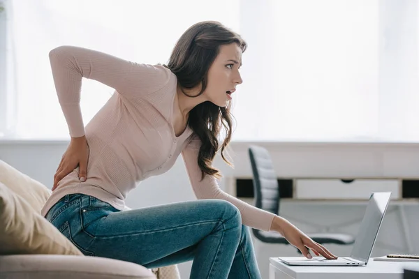 Side view of young woman suffering from back pain while using laptop at home — Stock Photo