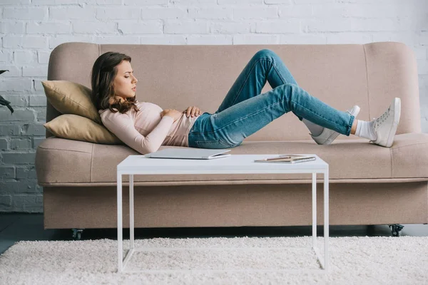 Vista laterale di giovane donna con dolore addominale sdraiata sul divano a casa — Foto stock