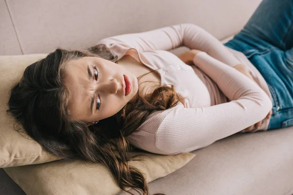 Junge Frau mit Bauchschmerzen auf Sofa liegend und zu Hause wegschauend — Stockfoto