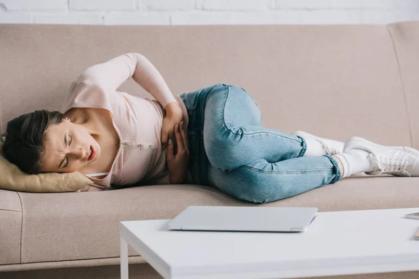 Young woman suffering from stomach ache while lying on sofa at home — Stock Photo