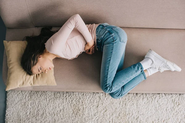 Vista superior da jovem mulher que sofre de dor de estômago enquanto deitada no sofá em casa — Fotografia de Stock