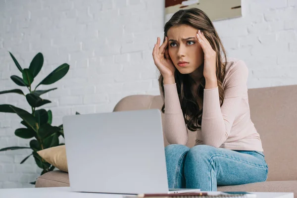 Mujer joven con dolor de cabeza mirando hacia otro lado mientras trabaja con el ordenador portátil en casa - foto de stock