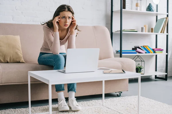 Mujer joven que sufre de dolor de cabeza y mirando hacia otro lado mientras usa el ordenador portátil en casa - foto de stock