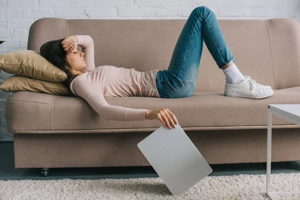 Vue latérale de la jeune femme avec des maux de tête couché sur le canapé et tenant ordinateur portable — Photo de stock