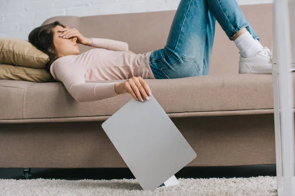 Молодая женщина с головной болью и болью в глазах лежит на диване и держит ноутбук — стоковое фото