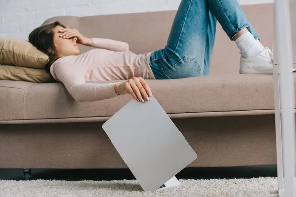 Mujer joven con dolor de cabeza y dolor en los ojos acostado en el sofá y la celebración de la computadora portátil - foto de stock